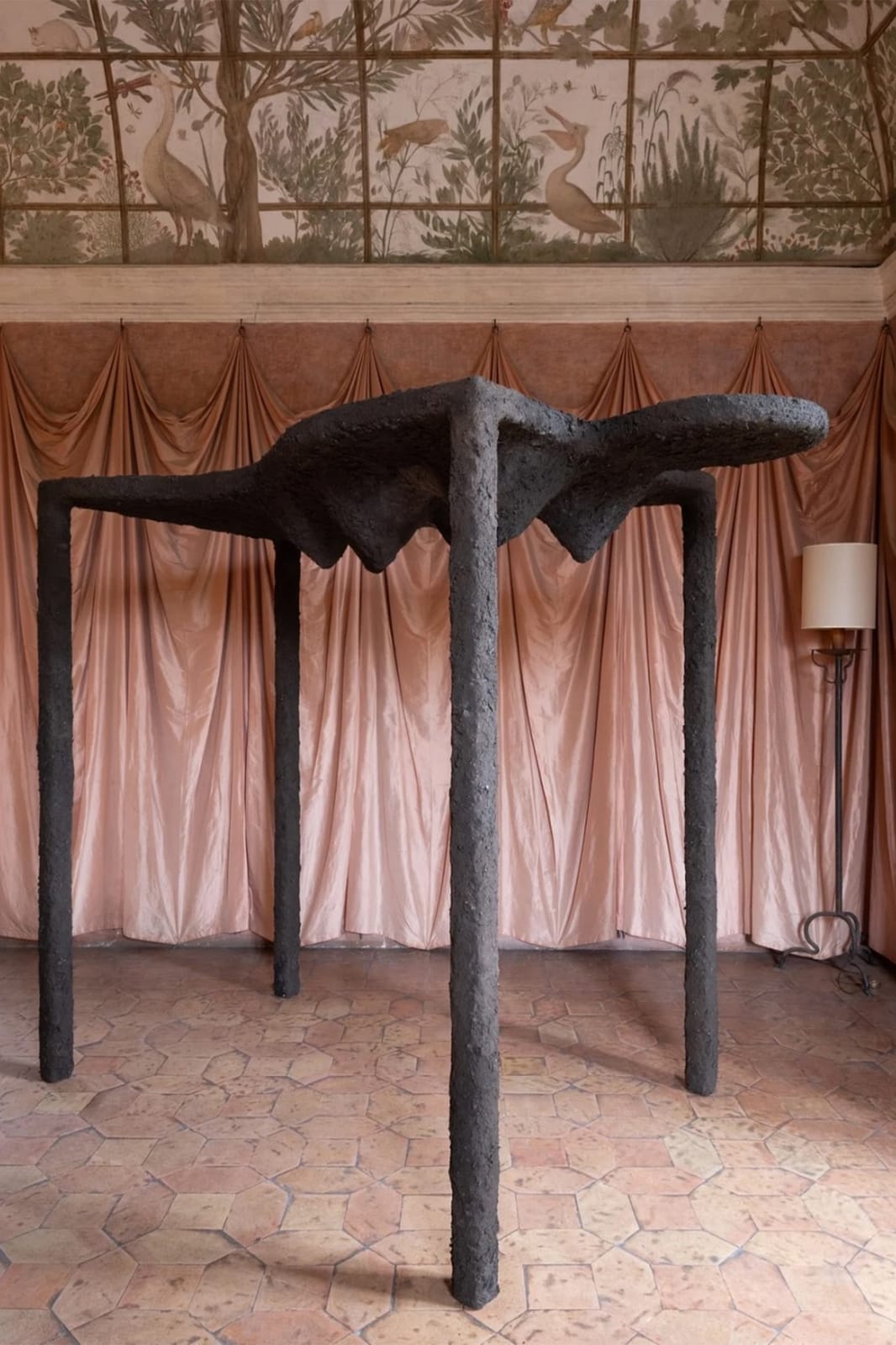 Bronze creature on four threadlike legs before a pink drapery, terracotta tiled floor