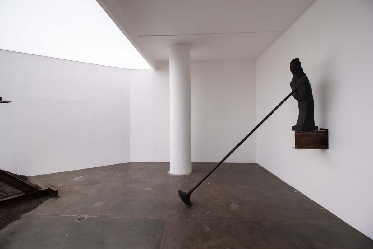 Bronze figure fixed to the wall, its threadlike leg reaching the concrete floor, São Paulo Biennial